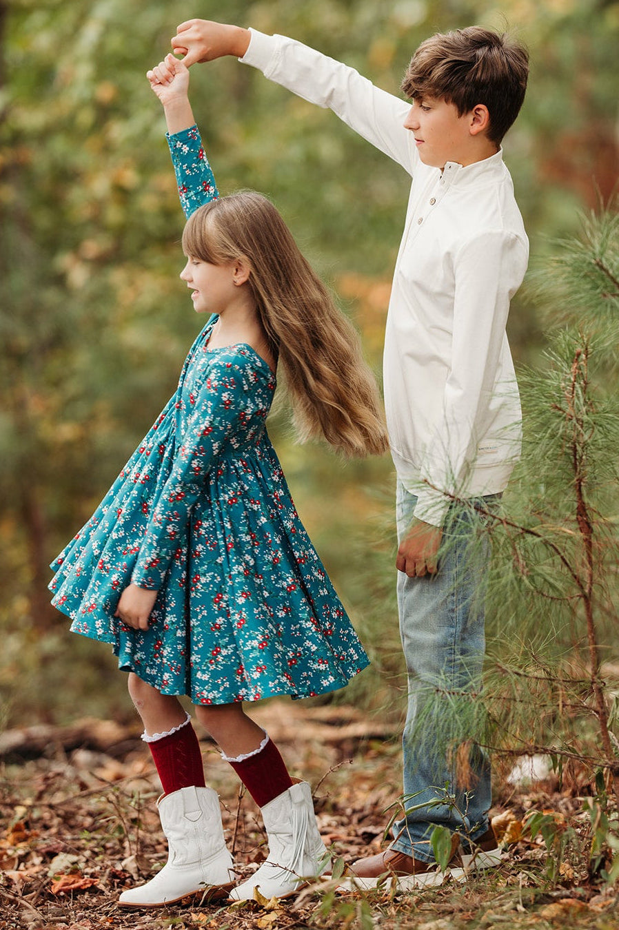 Two children, a girl in a teal holiday berry dress and white boots, and a boy in a cream pullover and jeans, standing in a forest.