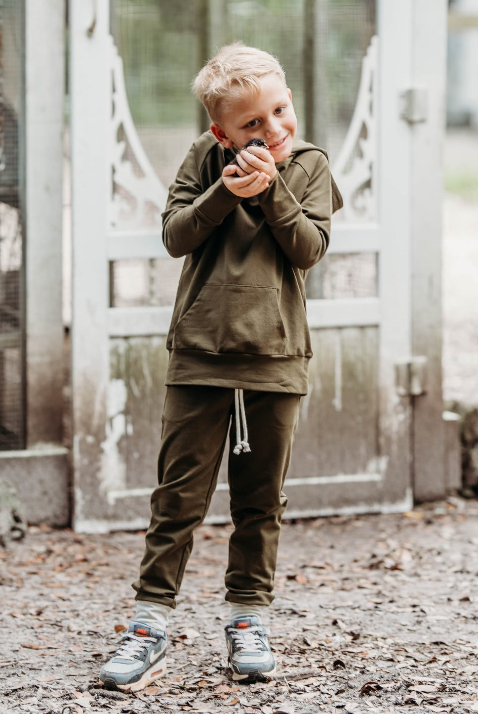 Child in an olive green hoodie and joggers standing in front of a door with a blurred outdoor background