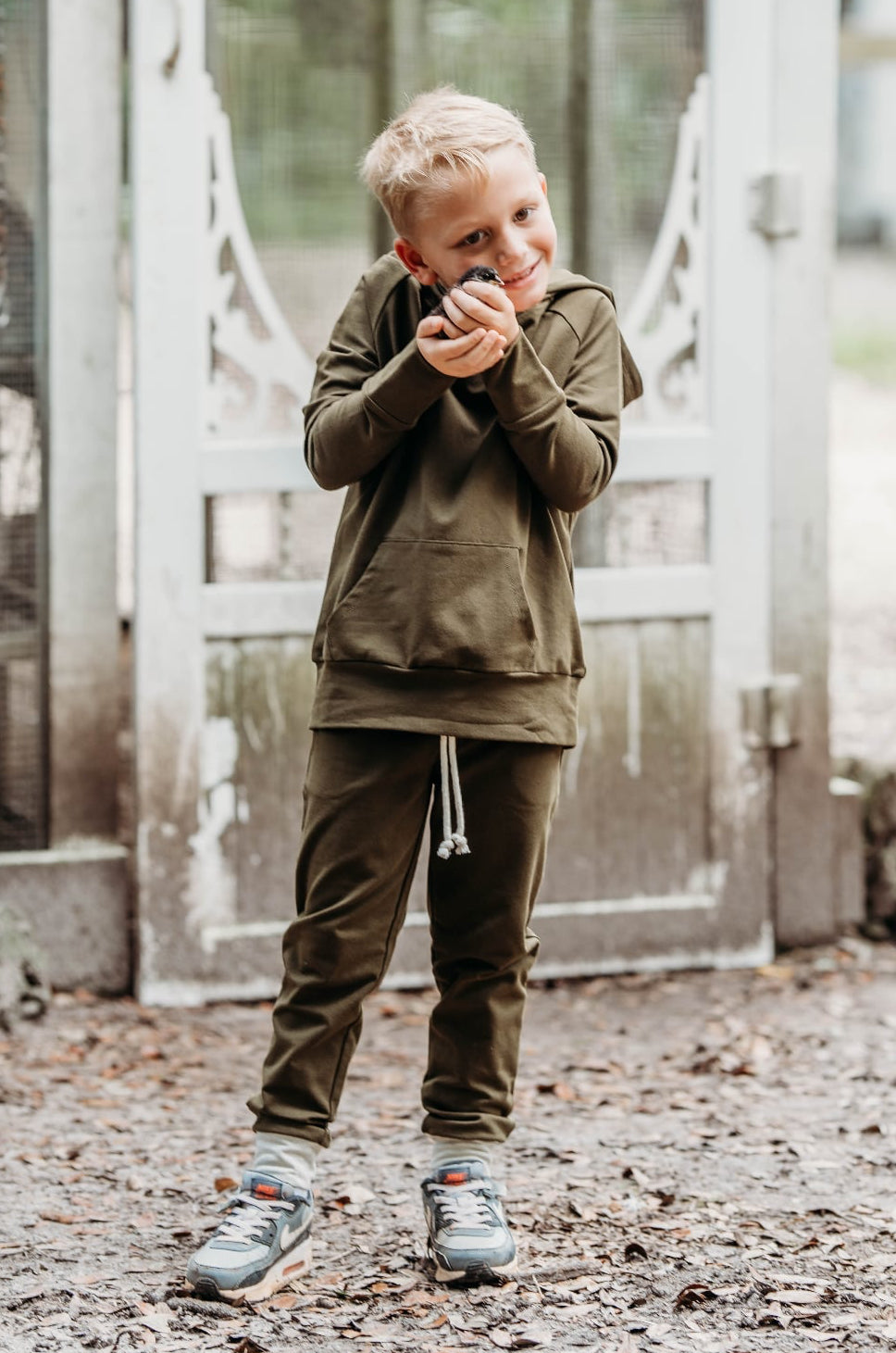 Child in an olive green hoodie and joggers standing in front of a door with a blurred outdoor background