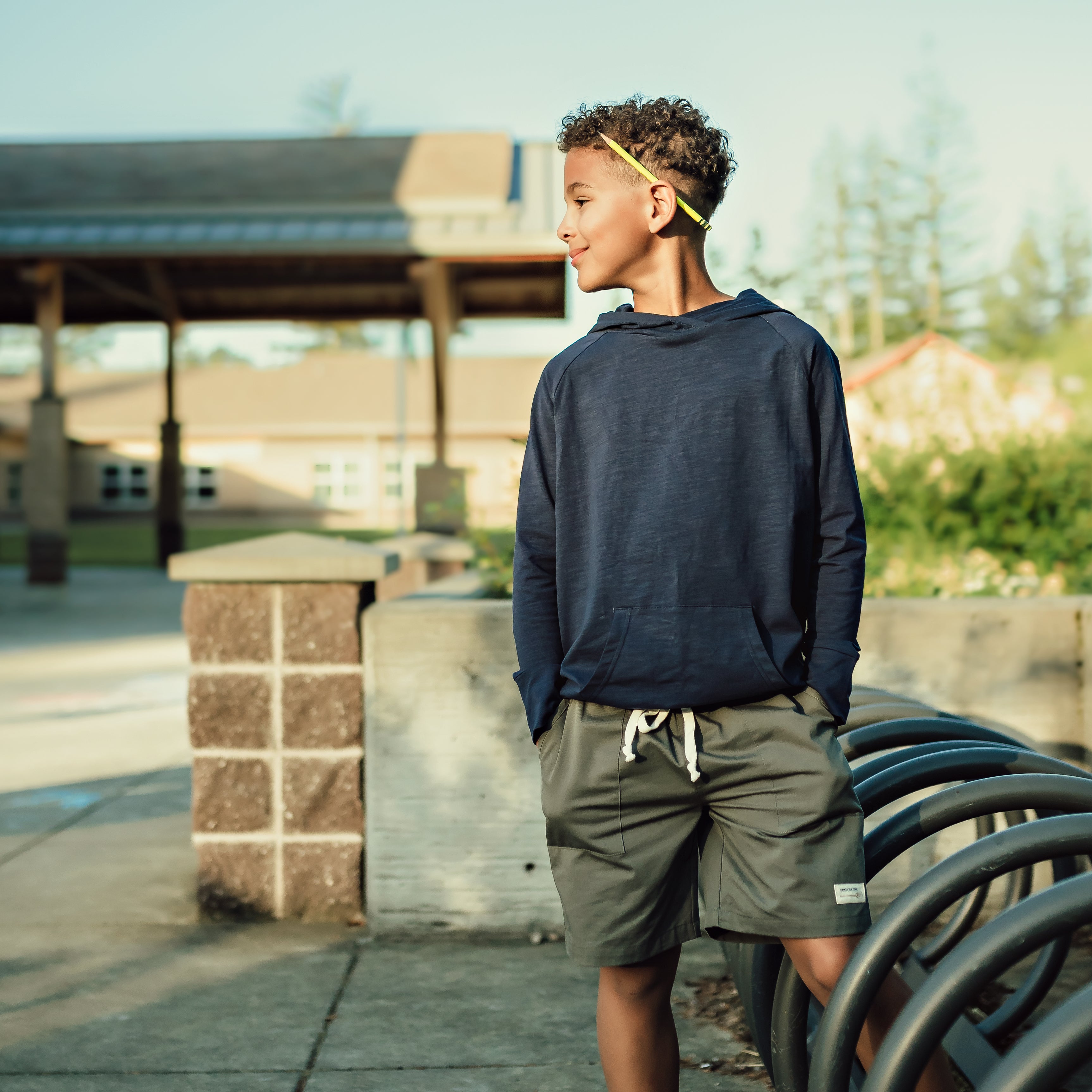 Boy standing near the school bike racks looking to the side with a pencil behind his ear. He is wearing a Sawyer & Finn navy hoodie and shorts.