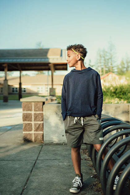 Boy standing near the school bike racks looking to the side with a pencil behind his ear. He is wearing a Sawyer & Finn navy hoodie and shorts.