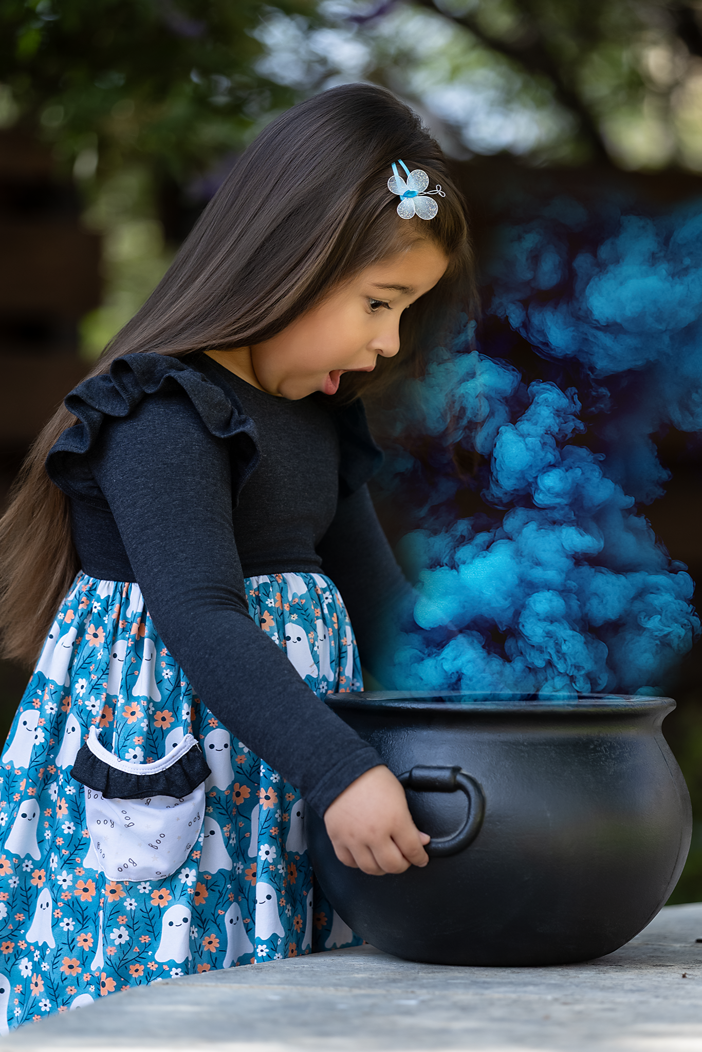 Child in Halloween dress with a blue smoke effect near a black cauldron outdoors