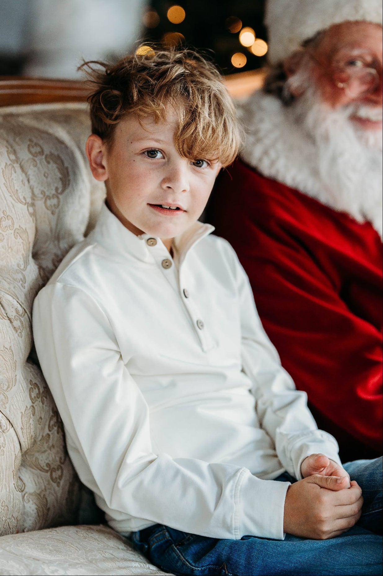 Young boy wearing a cream pullover sitting next to Santa Claus on a festive background